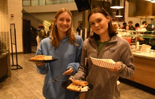 Two female students hold out plates of breakfast food in front of a dining hall station