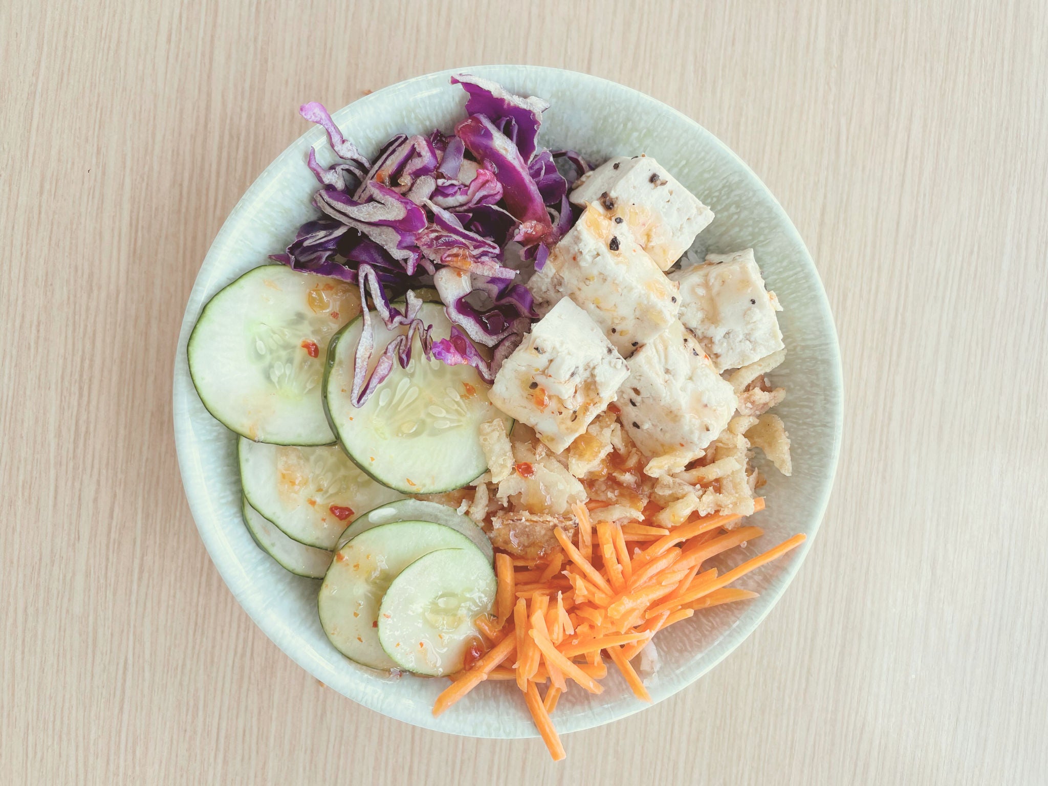 Overhead view of a bowl featuring cubed tofu seasoned with black pepper, served alongside sliced cucumber, shredded carrots, and shredded purple cabbage on a light wood surface.