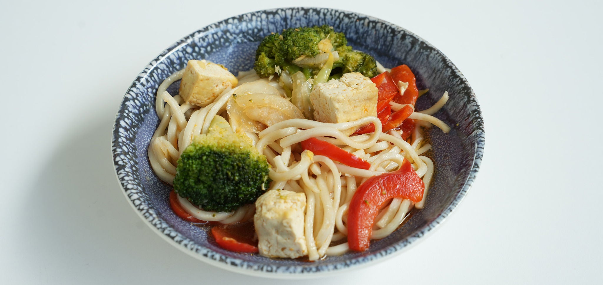 Tofu Noodle Bowl Hero Image.jpg: A top-down shot of a wide blue bowl filled with thick white noodles, cubed tofu, broccoli florets, and sliced red bell peppers in a light sauce