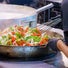 A chef in black gloves uses a large metal whisk to toss a colorful mixture of sliced green peppers, red peppers, and onions in a large stainless steel pan.