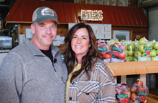 One male and one female farmer stand in their store in front of produce.