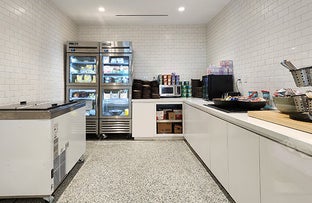 A clean, organized campus pantry with white tiled walls, featuring glass-door refrigerators, a microwave, and counters stocked with snack bins and supplies