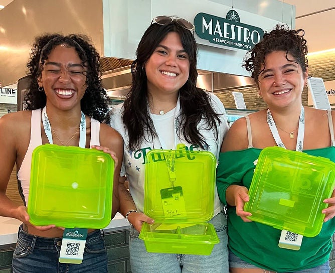 Three people smiling and holding up bright neon-green, reusable plastic to-go containers in the Malkin Sacks Commons.