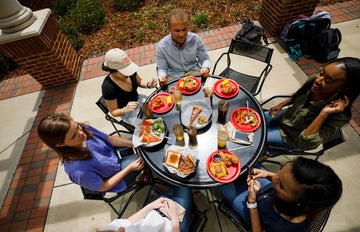 A group of college students sit around a round table with plates of food and drinks eating outside.