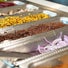 A close-up, angled view of a salad bar featuring metal trays filled with various fresh toppings, including roasted corn, red quinoa, sliced red onions, and shredded cheese.